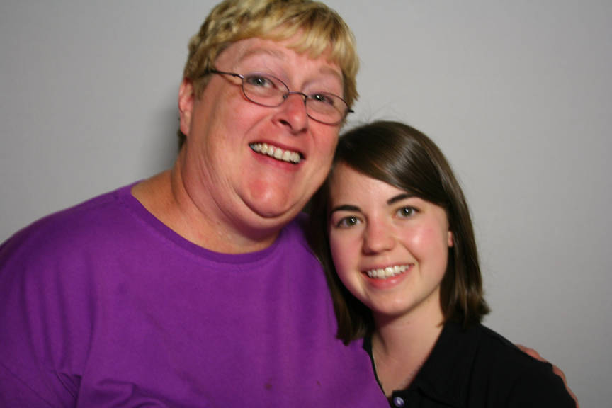 Two women smile for the camera, one with blonde hair, glasses, and a purple shirt, and the other with brown hair and a black shirt