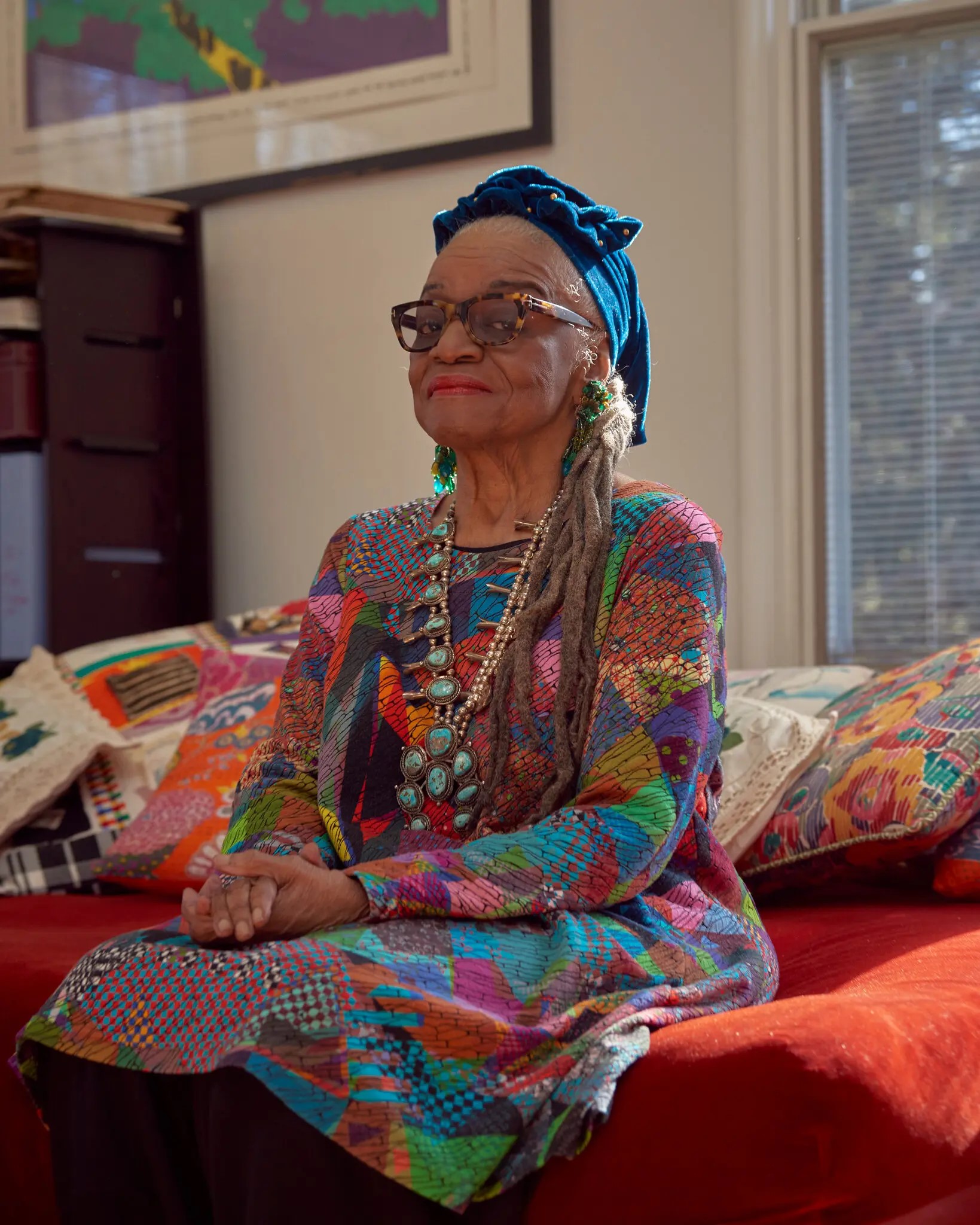 Image of artist Faith Ringgold. She is a slim older African American woman, seated on a red couch. She wears a printed dress, a blue head wrap, and glasses. 