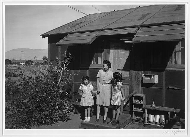 Photograph by Ansel Adams, 1943. Black and white photograph shows Mrs. Yaeko Nakamura and her two children, Joyce Yuki (right) and Louise Tami (left), standing on the step at the entrance of a wooden dwelling, Manzanar Relocation Center. 