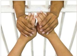 One pair of tanned hands holding another pair of tanned hands through white jail cell bars. 