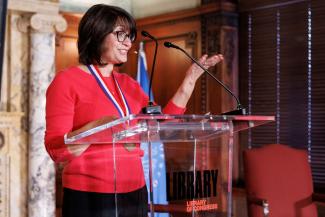 Author Meg Medina, wearing a red sweater, speaks at a plexiglass podium at the Library of Congress. Photo credit Shawn Miller.  