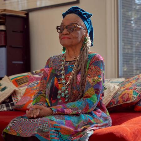 Image of artist Faith Ringgold. She is a slim older African American woman, seated on a red couch. She wears a printed dress, a blue head wrap, and glasses. 