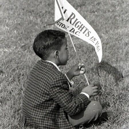 Black boy in suit sitting on grass holding Civil Rights March 1963 pennant flag